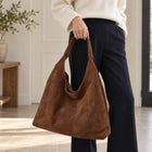 Coffee faux suede shoulder bag held by its single strap at the side